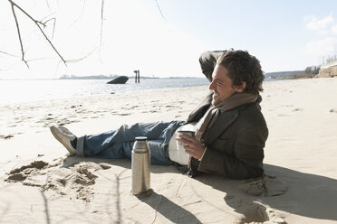 Germany, Hamburg, Man lying on sand with coffee flask near Elbe riverside - DBF000101