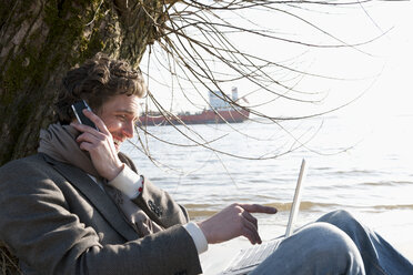 Germany, Hamburg, Man using laptop and cell phone near Elbe riverside with container ship in background - DBF000125