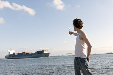 Germany, Hamburg, Man with beer bottle near Elbe riverside and container ship in background - DBF000130