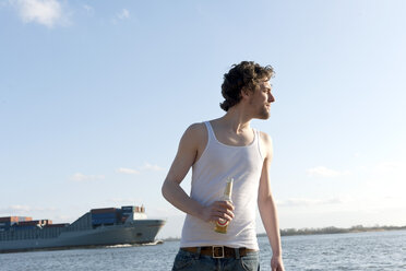 Germany, Hamburg, Man with beer bottle near Elbe riverside and container ship in background - DBF000131