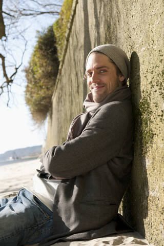 Germany, Hamburg, Mid adult man sitting and leaning against wall near Elbe river, smiling, portrait stock photo