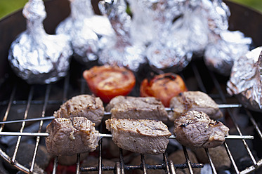 Germany, North Rhine-Westphalia, Düsseldorf, Close up of shashlik spears, tomatoes and onions in aluminium foil on grill - KJF000088