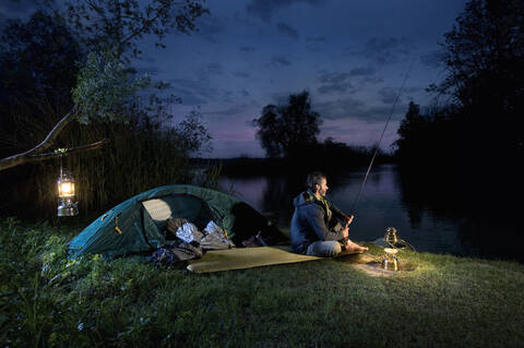 Germany, Bavaria, Ammersee, Man sitting near lakeshore with lantern and fishing at dusk stock photo