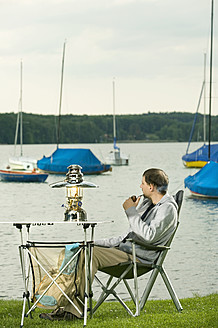 Germany, Bavaria, Woerthsee, Man smoking pipe near lakeshore while camping - RNF000611