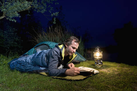 Germany, Bavaria, Ammersee, Man reading book near lakeshore while camping at night stock photo