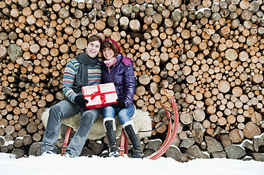 Österreich, Salzburger Land, Flachau, Junger Mann und Frau auf Schlitten sitzend mit Weihnachtsgeschenk und Brennholz im Hintergrund - HHF003629