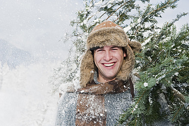 Austria, Salzburg Country, Flachau, Young man carrying christmas tree in snow - HHF003650
