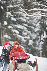 Österreich, Salzburger Land, Flachau, Junge Frau sitzt auf Schlitten mit Weihnachtspaketen und Mann schiebt ihn - HHF003677