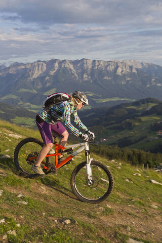 Österreich, Tirol, Spitzstein, Junge Frau beim Mountainbiken auf der Piste mit dem Kaisergebirge im Hintergrund, lizenzfreies Stockfoto