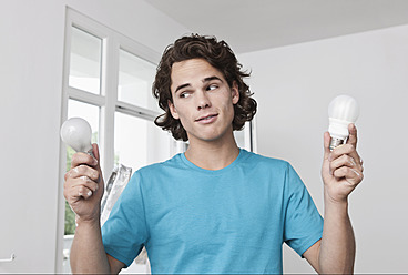 Germany, Cologne, Young man holding normal and energy saving lightbulbs in renovating apartment - FMKF000275