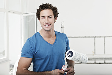 Germany, Cologne, Close up of young man holding paint roller in renovating apartment, portrait, smiling - FMKF000324