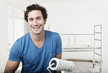 Germany, Cologne, Close up of young man holding paint roller in renovating apartment, portrait, smiling - FMKF000325