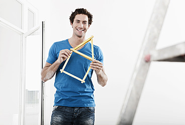 Germany, Cologne, Young man holding house model pocket ruler in renovating apartment - FMKF000333