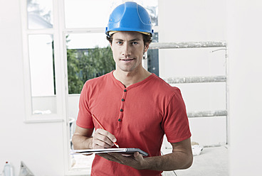 Germany, Cologne, Young man wearing hard hat writing on writing pad in renovating apartment - FMKF000334
