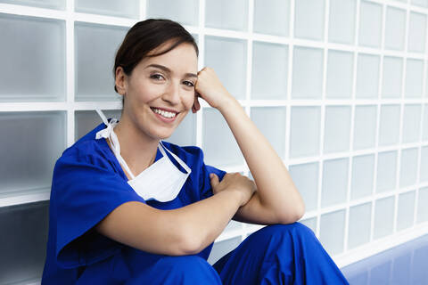 Germany, Bavaria, Diessen am Ammersee, Young doctor wearing scrubs and sitting in corridor, smiling, portrait stock photo