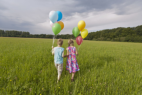 Deutschland, Nordrhein-Westfalen, Hennef, Mädchen und Junge halten Luftballons und gehen auf einer Wiese spazieren - KJF000128