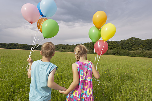 Germany, North Rhine-Westphalia, Hennef, Girl and boy holding balloons and walking in meadow - KJF000129