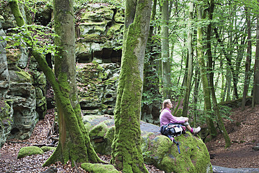 Germany, Rhineland-Palatinate, Eifel Region, South Eifel Nature Park, View of woman hiker sitting on bunter rock formations at beech tree forest - GWF001536