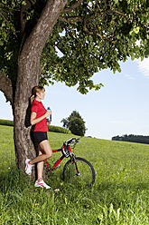 Germany, Bavaria, Young woman leaning against tree with mountain bike - MAEF003654