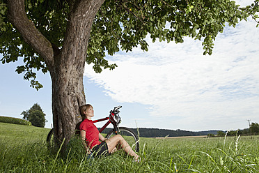 Germany, Bavaria, Young woman sitting against tree with mountain bike - MAEF003651