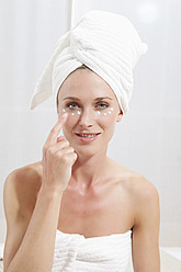 Junge Frau, eingewickelt in ein Handtuch, trägt Gesichtscreme auf - MAEF003678