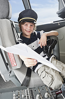 Germany, Bavaria, Munich, Boy wearing captain's hat and holding paper plane in airplane cockpit - WESTF017092