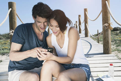Spanien, Mallorca, Junges Paar mit Kamera auf Strandpromenade sitzend, lächelnd, lizenzfreies Stockfoto