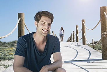 Spanien, Mallorca, Junger Mann sitzt auf Promenade und Frau geht im Hintergrund - WESTF017123