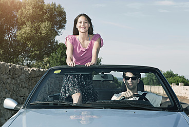 Spain, Majorca, Young man driving and woman standing in cabriolet car - WESTF017159