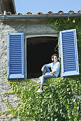 Italy, Tuscany, Young man sitting on window sill with shutters - PDF000183