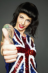 Young woman showing her thumb with tatto on her hand against grey background, portrait, smiling - MBE000188