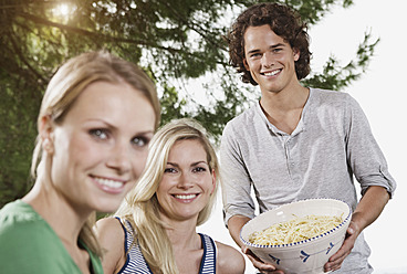 Italy, Tuscany, Magliano, Young man holding bowl of pasta and women in foreground, smiling, portrait - WESTF017374