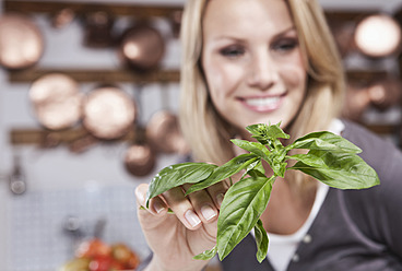Italy, Tuscany, Magliano, Young woman holding basil leaf in kitchen, smiling - WESTF017422