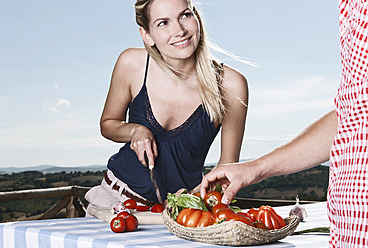 Italien, Toskana, Magliano, Junger Mann steht bei Tisch und Frau schneidet Tomaten, lächelnd - WESTF017449