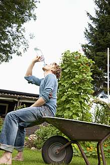 Deutschland, Hamburg, Mann sitzt auf Schubkarre und trinkt Wasser in Kleingartenanlage - DBF000180