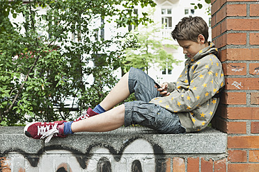Germany, Berlin, Boy sitting against brick wall with cell phone - WESTF017481