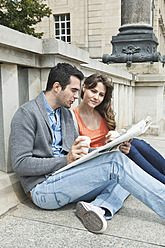 Germany, Berlin, Couple sitting on sidewalk and reading newspaper - WESTF017564