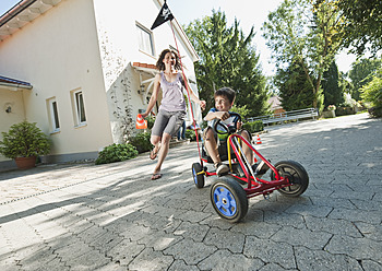 Germany, Bavaria, Boy driving pedal go kart and woman running with family in background - WESTF017811