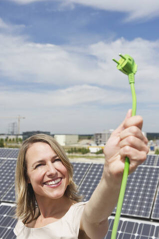 Deutschland, München, Frau hält Stromkabel in Solarenergie, lächelnd, lizenzfreies Stockfoto