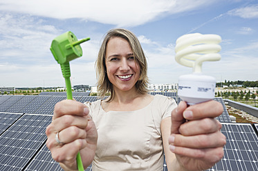 Deutschland, München, Frau hält Stromkabel und Glühbirne in Solaranlage, lächelnd, Porträt - WESTF017836