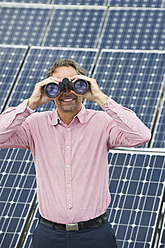 Germany, Munich, Mature man looking through binocular in solar plant, smiling - WESTF017857