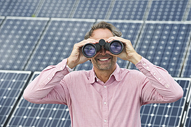 Germany, Munich, Mature man looking through binocular in solar plant, smiling - WESTF017858