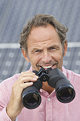 Germany, Munich, Mature man holding binocular in solar plant, smiling, portrait - WESTF017859