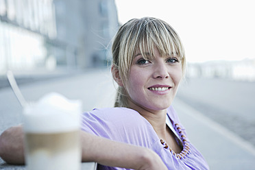 A cheerful young woman enjoys her coffee in the foreground, with the city of Cologne, Germany in the background - WESTF017950