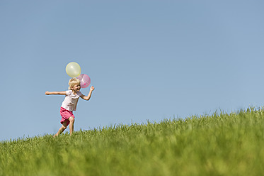 Eine fröhliche junge Frau sprintet über eine malerische bayerische Wiese, in der Hand einen bunten Strauß Luftballons und grinst von einem Ohr zum anderen - RNF000737