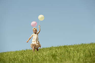 Ein fröhliches Mädchen sprintet durch eine malerische bayerische Wiese, hält einen bunten Strauß Luftballons in der Hand und grinst von einem Ohr zum anderen - RNF000738