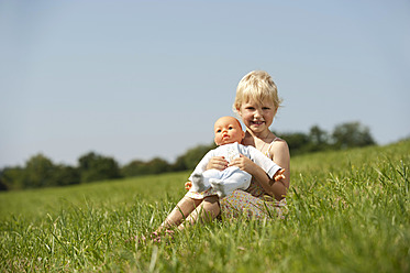 A happy Bavarian girl enjoys the sunshine with her doll, smiling in a beautiful green field - RNF000747