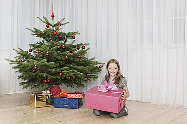 Fröhliches Mädchen posiert mit einem Geschenk unter dem Weihnachtsbaum in München, Deutschland, und strahlt vor Freude in einem Porträt - SKF000629