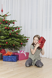 Ein junges Mädchen wartet sehnsüchtig auf den Weihnachtsmorgen und hält ein schön verpacktes Geschenk in der Nähe eines festlich geschmückten Baumes in München, Deutschland - SKF000630