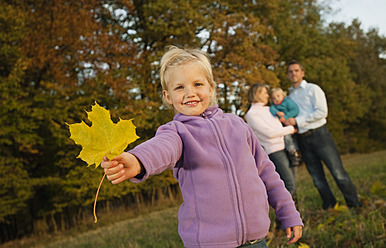 Ein junges Mädchen bewundert ein buntes Herbstblatt, während ihre Familie im Hintergrund steht, in Bayern - RNF000832